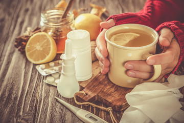 Medical care concept - hands holding cup with ginger honey and lemon tea with drugs, pills and spray on wood background