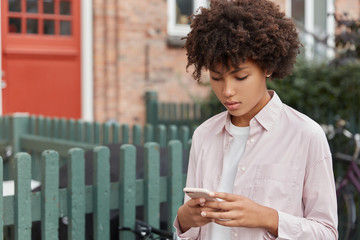 Horizontal view of serious concentrated cute Afro girl in stylish shirt, holds modern smart phone,...