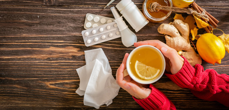 Medical care concept - hands holding cup with ginger honey and lemon tea with drugs, pills and spray on wood background