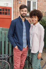 Vertical shot of cheerful mixed race couple have date, stand closely to each other near bicycles,...
