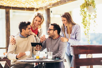 Group of four friends having fun a coffee together. Two women and two men at cafe talking laughing and using digital tablet.