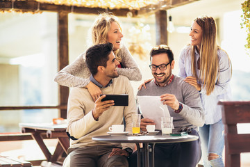 Group of four friends having fun a coffee together. Two women and two men at cafe talking laughing and using digital tablet.