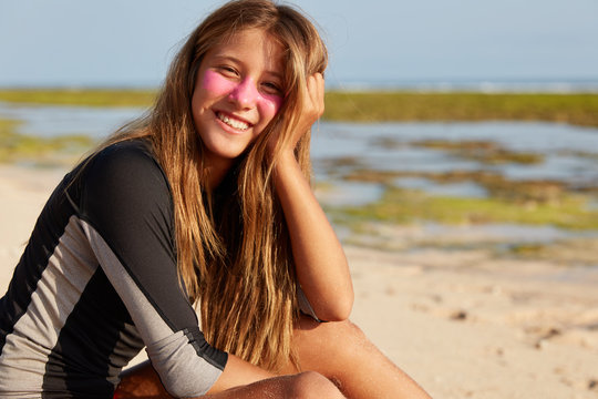 Summer time, resort and positive emotions concept. Beautiful long haired woman with cheerful expression, has surf zic mask, relaxes on beach, dressed in black wetsuit, copy space for your promotion