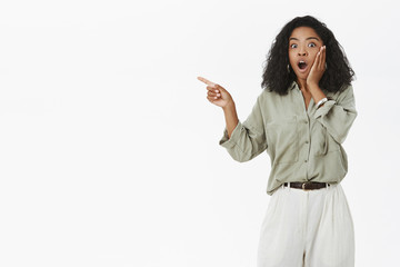 Portrait of impressed speechless and astonished young dark-skinned female fan in stylish blouse with curly hair saying wow touching cheek from amazement pointing left with excitement