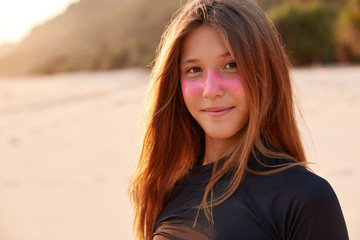 Close up shot of beautiful Caucasian girl with pink zurf zinc mask, dressed in black wetsuit, enjoys sunshine, looks happily directly at camera, has recreation time in exotic paradise country