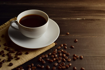 Black coffee in a white cup on hemp sack with coffee beans all around on top of black wooden floor in warm vintage tone with copy space for text, morning fresh up concept