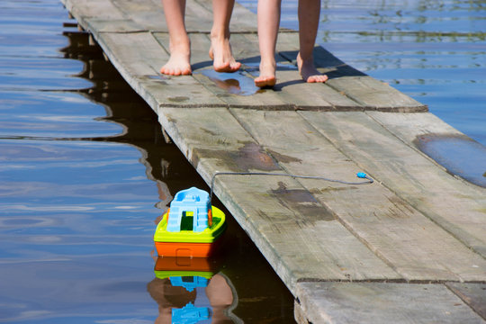two pairs of bare feet walking on the wooden laying to the toy boat in the lake