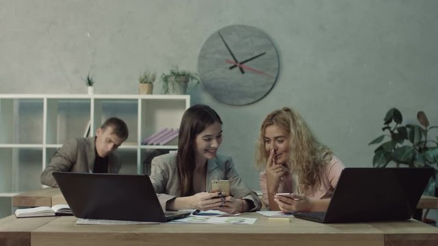 Inefficient lazy female office workers with cellpones distracting from work during business hours. Chatting office women browsing social media on smart phone and caught by angry boss at workplace.