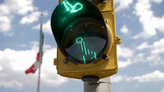 Streetlight Crosswalk Sign with Little Green Running Man in Front of Mexico City's Main Mexican Flag in the Center of the City(Zocalo). Blue Sky with Clouds Appear in Background
