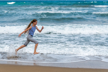 Cute happy little girl running along the beach in jumping over waves. Beautiful summer sunny day, blue sea, picturesque landscape.