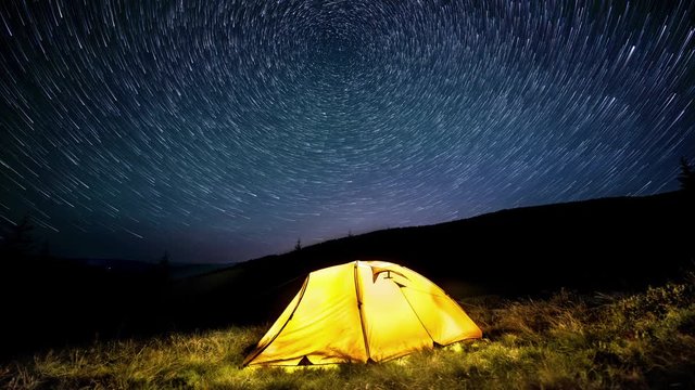 Time-lapse. Star circles above the night mountains and a glowing camping tent