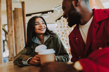 Sharing earphones. Cheerful lovely young couple sharing earphones and listening to pleasant music while sitting together and drinking coffee