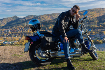 Obraz premium Young man wearing a black leather jacket, sunglasses and jeans sits outdoor on a motorcycle, resting on a mountain above the river. Lifestyle, travel. Copy space.