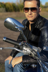 Fototapeta na wymiar Young man wearing a black leather jacket, sunglasses and jeans sits outdoor on a motorcycle, on a mountain on a blue sky background. Lifestyle, travel. Copy space