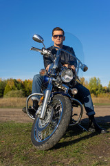 Fototapeta na wymiar Young man wearing a black leather jacket, sunglasses and jeans sits outdoor on a motorcycle, on a mountain on a blue sky background. Lifestyle, travel. Copy space
