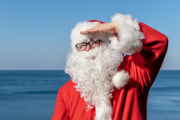 Santa looks into the distance, standing on the ocean. Traditional red outfit and relaxing on the beach