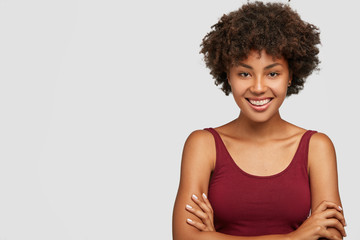 Friendly looking self assured dark skinned young girl keeps arms folded, has toothy smile, dressed in casual clothes, models against white background with free space for your promotional content