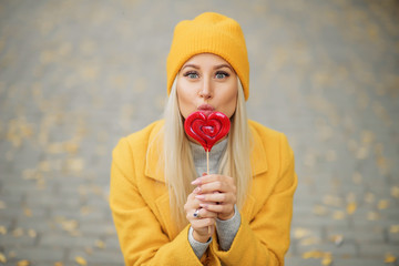 Fashion portrait pretty sweet young woman in yellow coat having fun with red lollipop heart over...