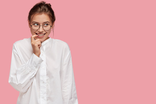 Photo of mysterious glad dark haired girl keeps index finger, looks positively aside, wears white shirt, poses against pink background with free space for your advertising content. Well what is there?