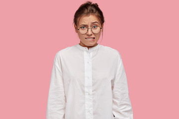 Doubtful nervous woman bites lips with embarrassed expression, raises eyebrows, has to make serious decision, wears casual white shirt, spectacles, models against pink background. Facial expressions