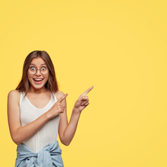 Photo of interested happy woman looks and points at upper right corner, feels awesome, poses over yellow background, dressed in casual clothes, shows nice shop, indicates at direction. Vertical shot