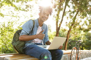 Fototapeta premium Emotional young african guy in park outdoors using laptop computer.
