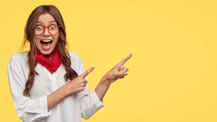 Poster Amazed attractive student indicates with both index fingers aside, dressed in white shirt, poses over yellow background, gazes at camera, keeps mouth opened, wears fashionable clothes. Wow, look there © Wayhome Studio