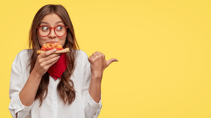 Photo of attractive woman eats slice of pizza, points aside with thumb, dressed in fashionable...