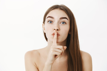 Naklejka premium Close-up shot of tender and sensual feminine brunette female posing naked showing shhh gesture asking keep secret of beauty safe standing calm and soft over gray background hiding surprise