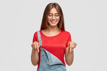 Horizontal shot of pleasant looking woman with cheerful expression, points don with both index...