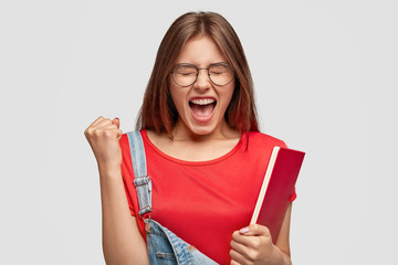 Angry nervous woman yells with annoyance, clenches fist, keeps mouth widely opened, expresses her anger, holds textbook, feels irritated as failed important exam, cant enter desirabale uniersity - obrazy, fototapety, plakaty