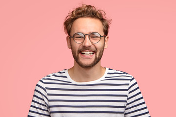 Headshot of pleased hipster has satisfied expression, curly hair and bristle, wears round...