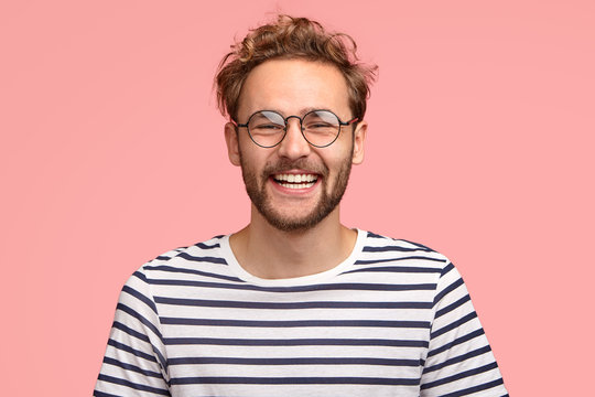Headshot of pleased hipster has satisfied expression, curly hair and bristle, wears round transparent glasses and striped t shirt, feels glad after promotion at work, isolated over pink background