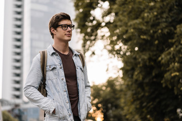 Young handsome tourist traveler with backpack walking on the street. Portrait of positive confident man with smiling face wearing blue jeans jacket and stylish hipster eyeglasses waiting for taxi. 