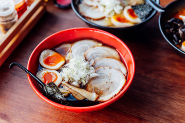 Shoyu Chashu Ramen: Japanese ramen in Shoyu sauce soup with chashu pork, boiled egg, dry seaweed and chives in red bowl on wooden counter.