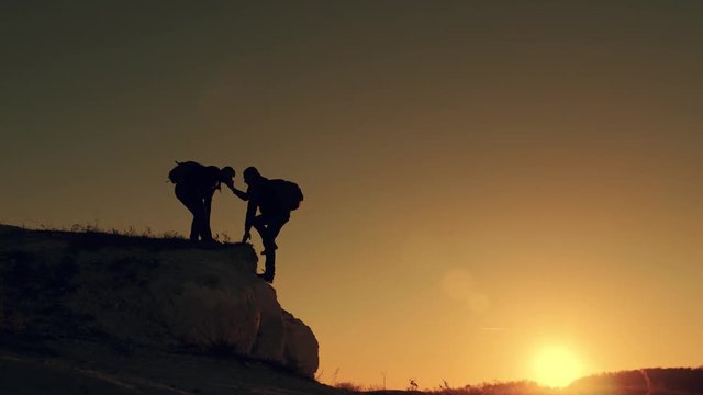 Silhouette of helping hand between two climber. two hikers on top of the mountain, a man helps a man to climb a sheer stone. couple hiking help each other silhouette in mountains with sunlight.
