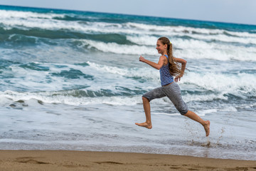 Fototapeta na wymiar Cute happy Girl in striped t-shirt running along the beach in jumping over waves. Beautiful summer sunny day, blue sea, picturesque landscape.