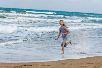 Fototapeta premium Cute happy Girl in striped t-shirt running along the beach in jumping over waves. Beautiful summer sunny day, blue sea, picturesque landscape.