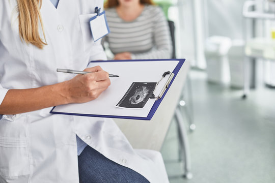 Close up of doctor hands holding pen and medical form of pregnant woman with sonography image of her child. Cropped woman sitting at the table on blurred background