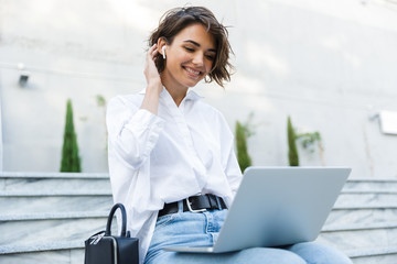 Beautiful woman sitting outdoors using laptop computer listening music with earphones.