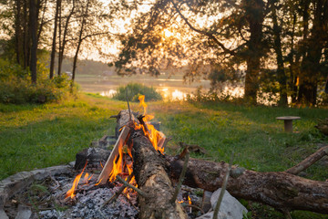 Lake side campfire with wood burning in a scenic setting during the sunset. Sun shines through canopy trees in golden light.
