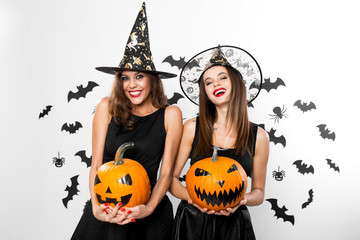 Two brunette girls in black dresses and witch`s hats hold horror halloween pumpkins on the background with bats