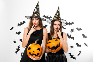 Two brunette girls in black dresses and witch`s hats hold horror halloween pumpkins on the background with bats