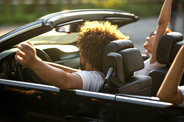 Curly dark-haired young man sitting at the wheel of a black cabriolet and his friends are sitting...