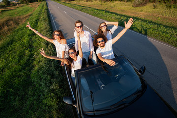 Stylish young guys are sitting and smiling in a black cabriolet on the country road on a sunny day.