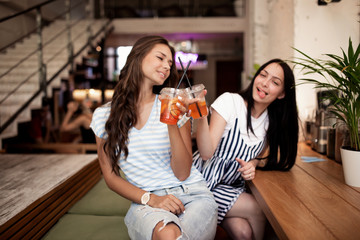 Two pretty youthful smiling girls,dressed in casual outfit,sit next to each other and look at the camera in a cozy coffee shop.