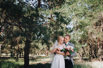 Beautiful newlyweds are standing in the woods and hugging. Wedding portrait of a stylish bride and cute bride with a large bouquet.