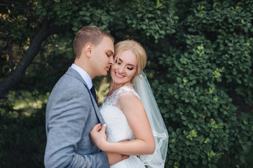 Obraz premium Beautiful newlyweds are smiling and hugging in a park with green trees. Wedding portrait of a young groom and cute bride. Wedding photography.