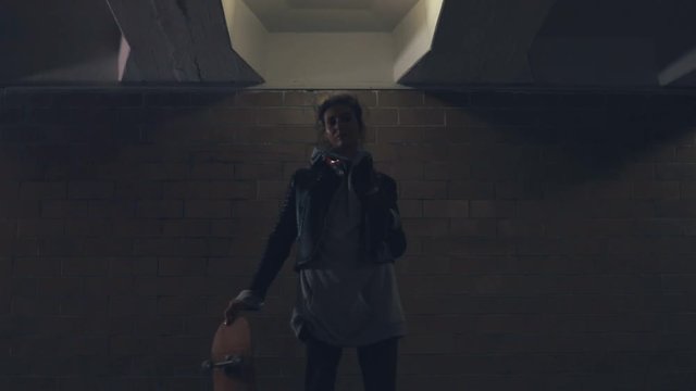 Girl standing in underpass at night. Young caucasian woman with skateboard in dark underground tunnel. Urban city lifestyle. Slow motion video shooting by handheld gimbal