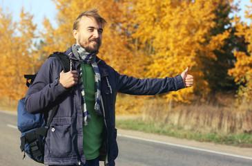 Young bearded hipster hitchhiker with backpack try to catch car on autumn road. Adventure travel in fall